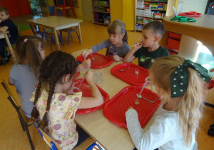Dzieci siedzą przy stoliku, w ręce trzymają pipetki, które napełniają wodą utlenioną i przelewają do pojemników z drożdżami.
